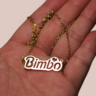 A close up of the Bimbo necklace from House Of Chastity being held in the palm of a hand, you can see that the pendant is in gold, with the lettering cut out.