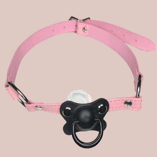 A pink pvc long gag that has a pale black  dummy fixed to the front which the wearer will have to hold in their mouth and a silver buckle fastening to the back. 